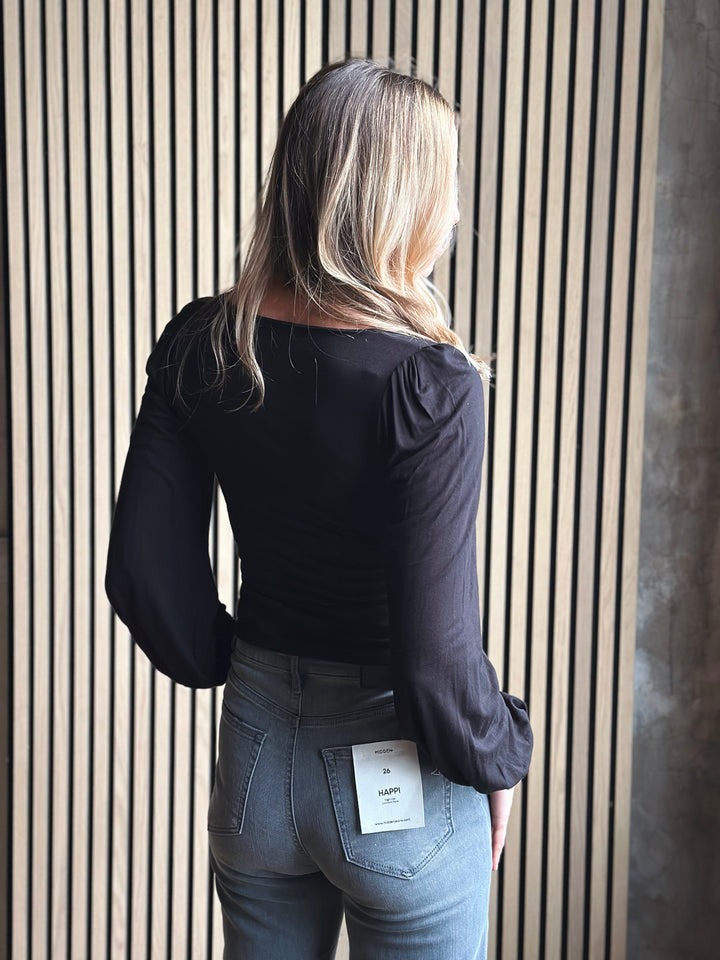 Person wearing a black top and gray jeans with a visible brand label, standing against a striped wall.