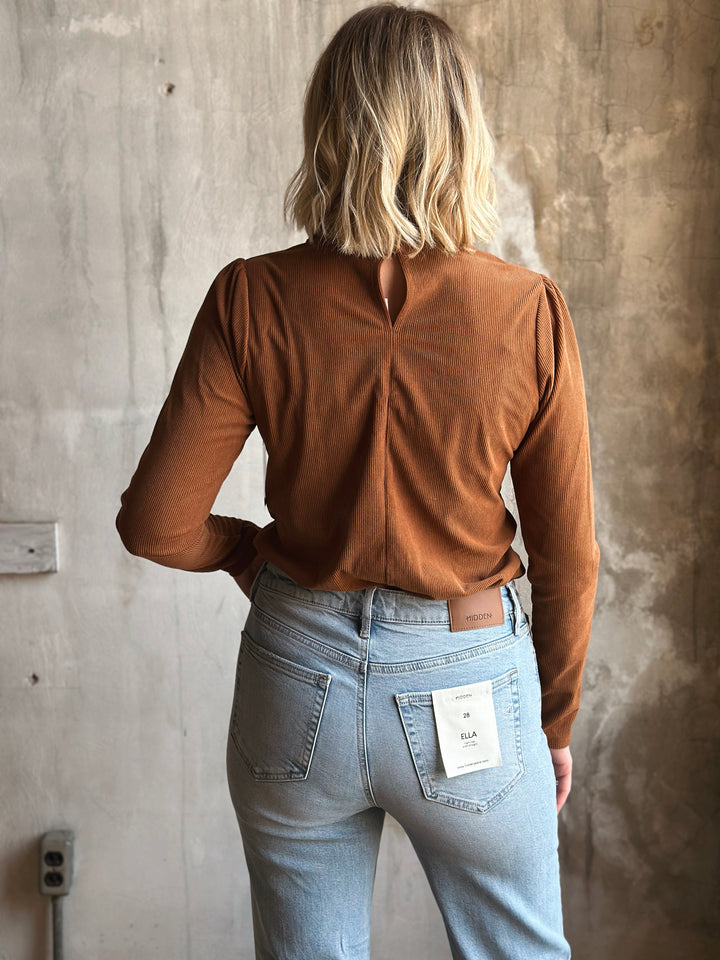 Person wearing a brown long-sleeve top and light blue jeans against a textured wall.