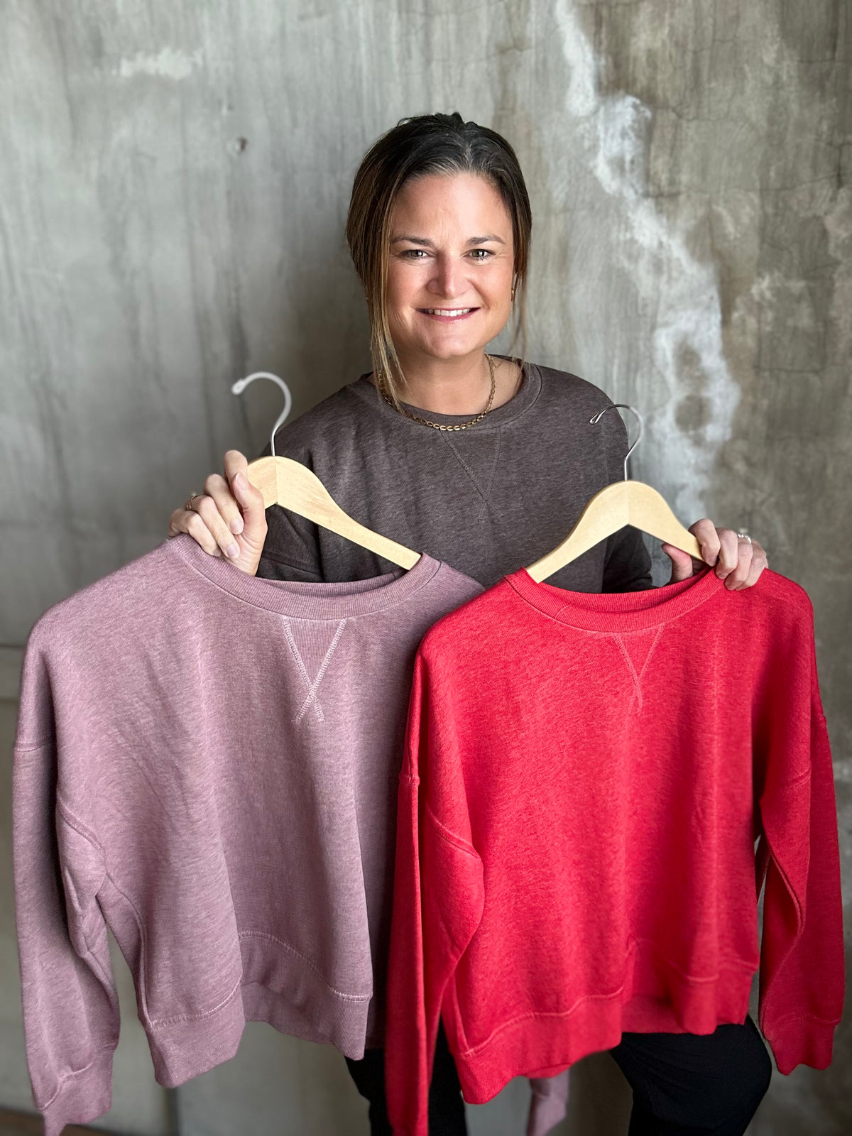 A woman holding up two crewneck sweatshirts on wooden hangers against a textured gray wall. The sweatshirt on the left is a dusty mauve-pink color, and the sweatshirt on the right is a bright red.