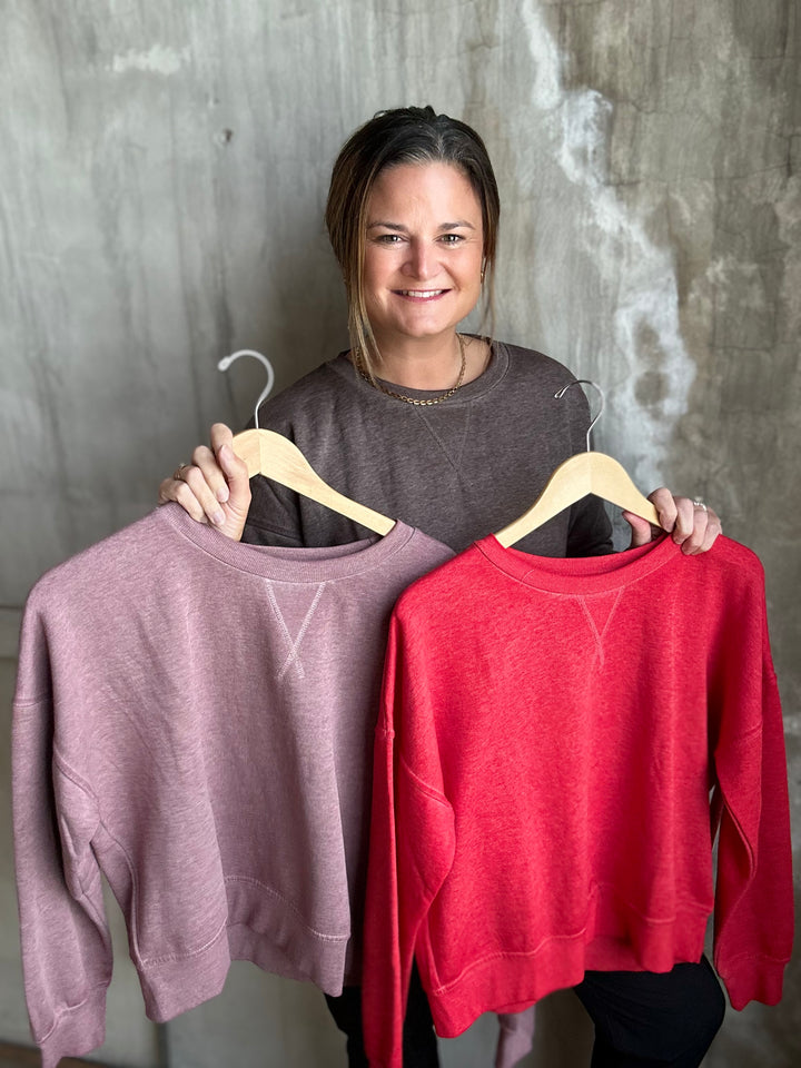 A woman holding up two crewneck sweatshirts on wooden hangers against a textured gray wall. The sweatshirt on the left is a dusty mauve-pink color, and the sweatshirt on the right is a bright red.