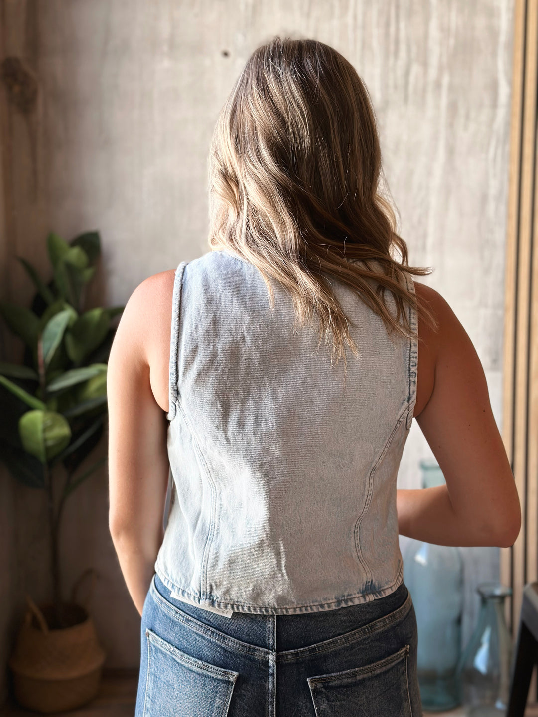 Person wearing a sleeveless denim top and jeans standing in a room with a plant and mirror.