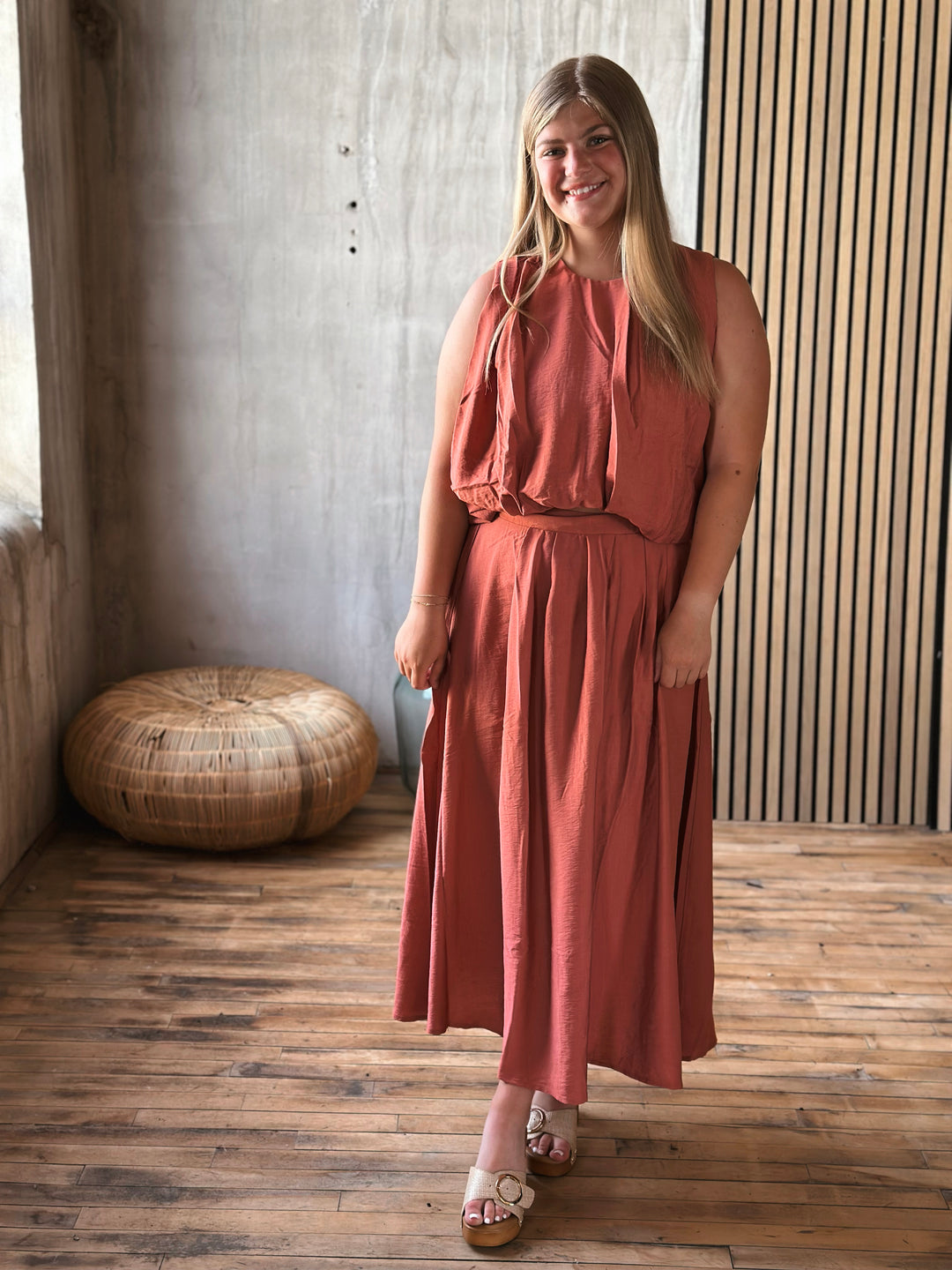 Woman in a rust-colored skirt and a sleeveless top set standing in a room with wooden flooring and a textured wall.