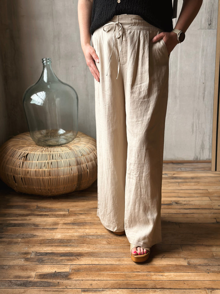 Person wearing beige linen pants standing in a room with wooden flooring and a large glass bottle.