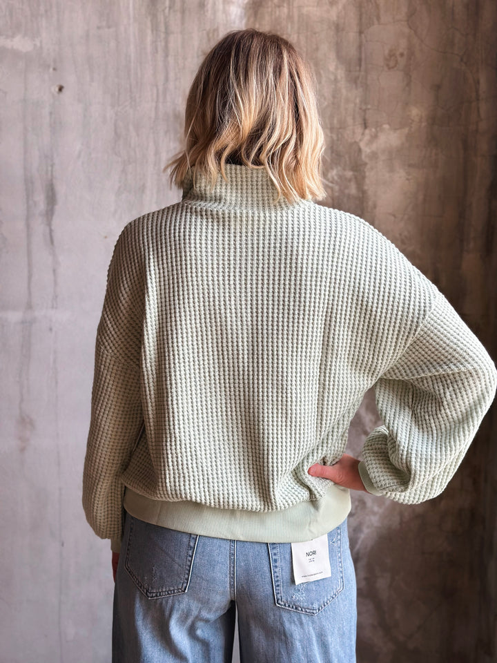 Person wearing a textured sweater and jeans against a concrete wall.