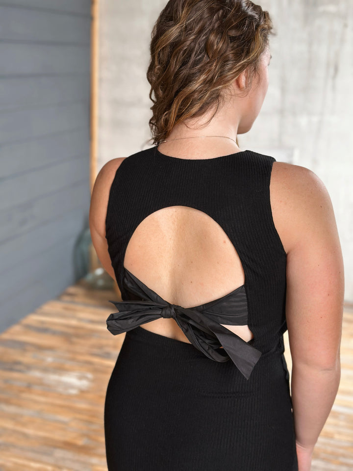 Woman wearing a black dress with an open back and bow detail, standing against a neutral background.