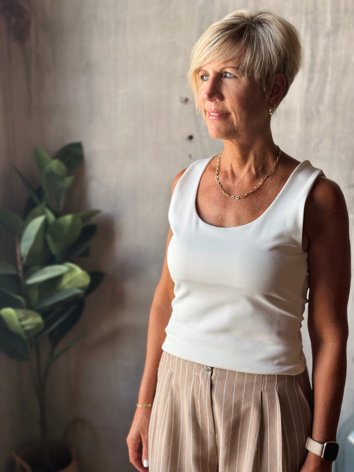 Woman wearing a white tank top and beige pants standing against a neutral wall with a plant in the background.