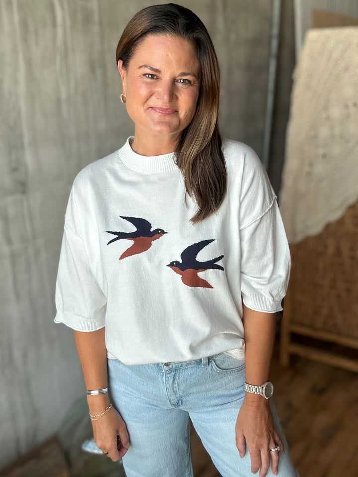 A woman wearing a white sweater with two embroidered flying swallow birds on the front, paired with light blue jeans.