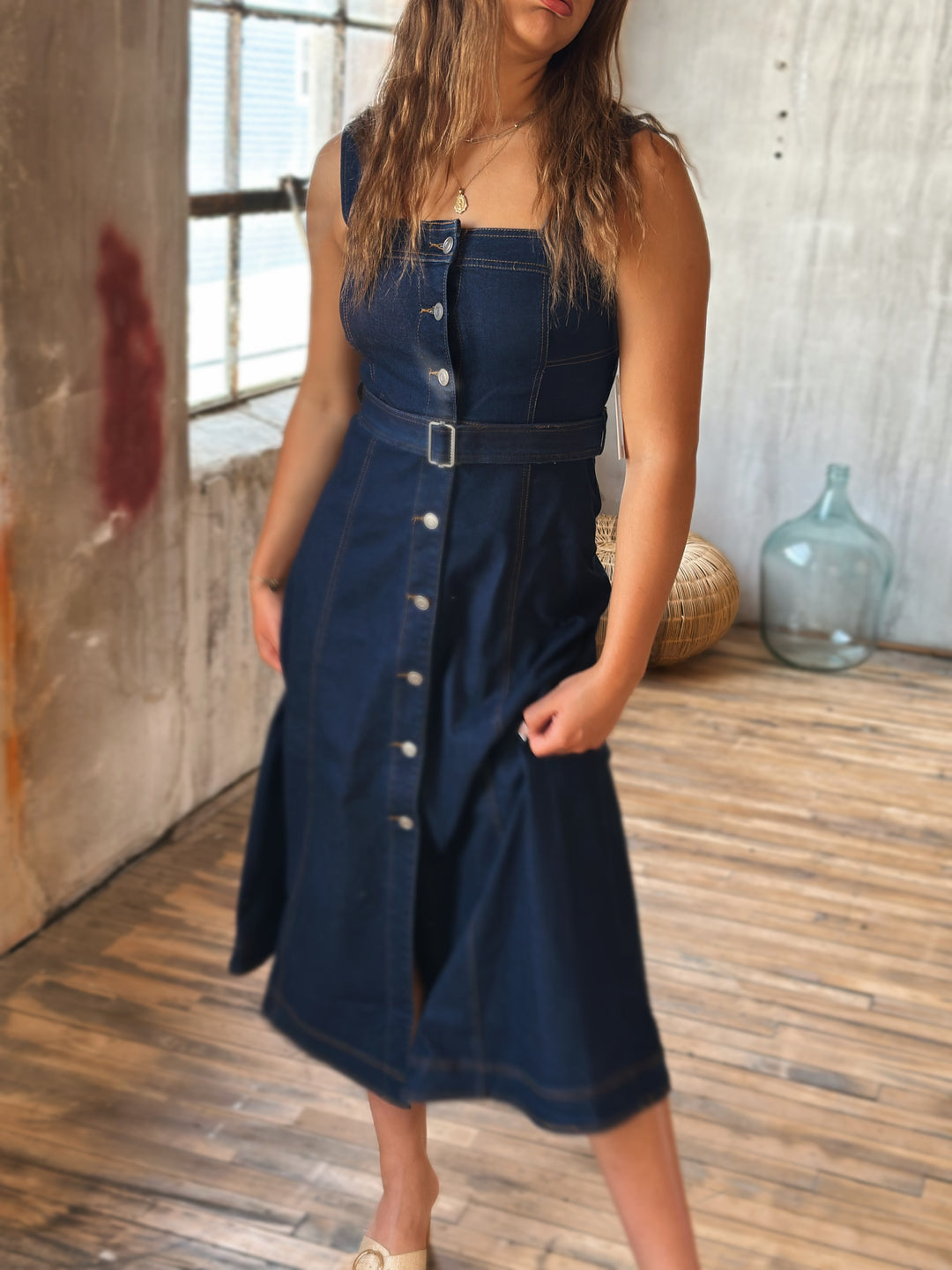 Woman wearing a dark blue denim dress with silver buttons in a room with wooden floor and textured walls.