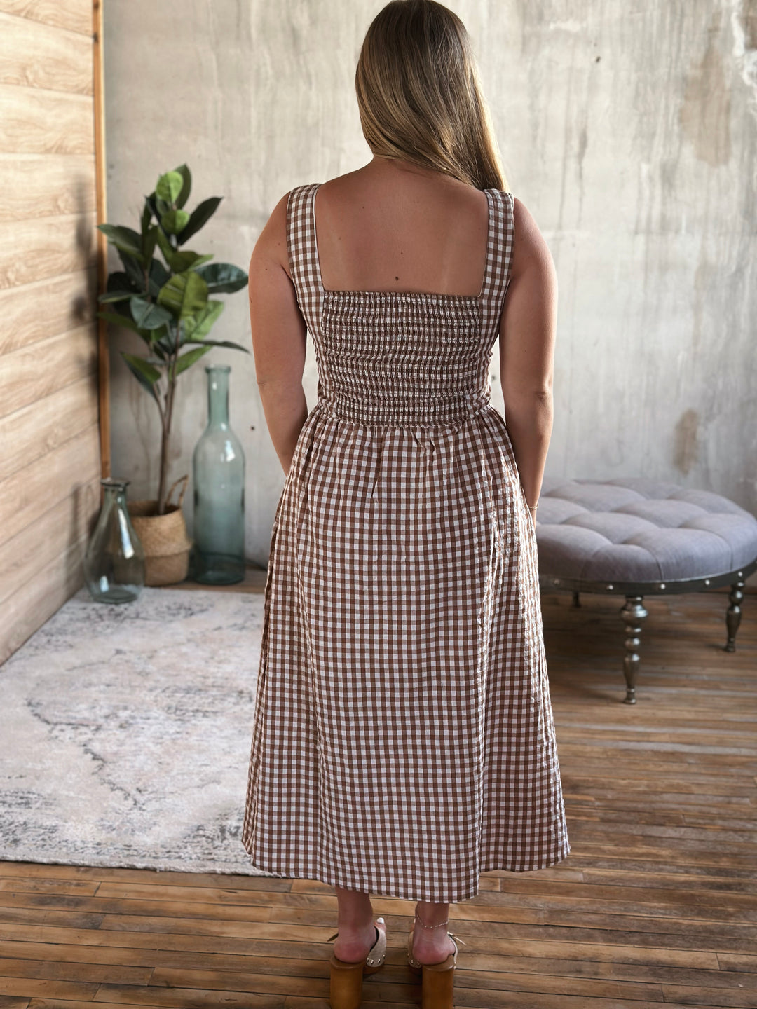 Woman wearing a brown and white plaid dress standing in a room with wooden floor and plant decor.