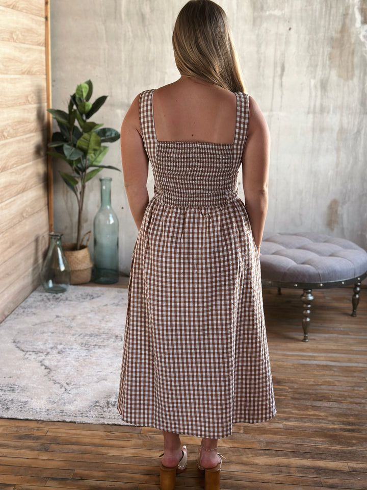 Woman wearing a brown and white plaid dress standing in a room with wooden floor and plant decor.