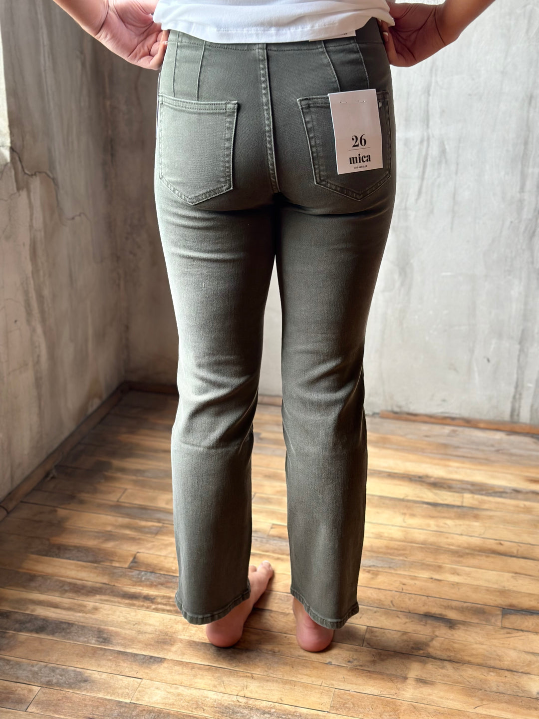 A person shown from the back wearing olive green jeans, standing barefoot on a wooden floor against a concrete wall. A white tag on the back pocket reads '26 mica'.