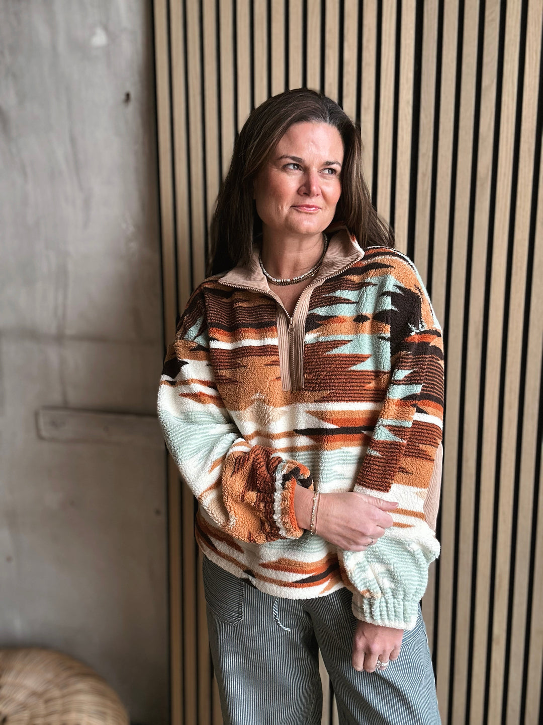 Woman wearing a patterned sweater standing against a wooden paneled wall.
