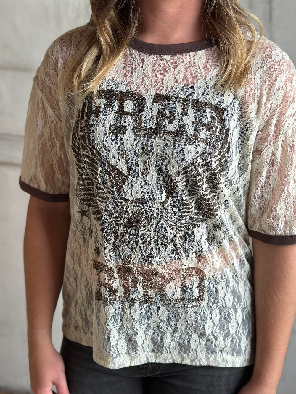 Close-up of a cream lace tee with a Free Bird eagle graphic and brown ringer trim