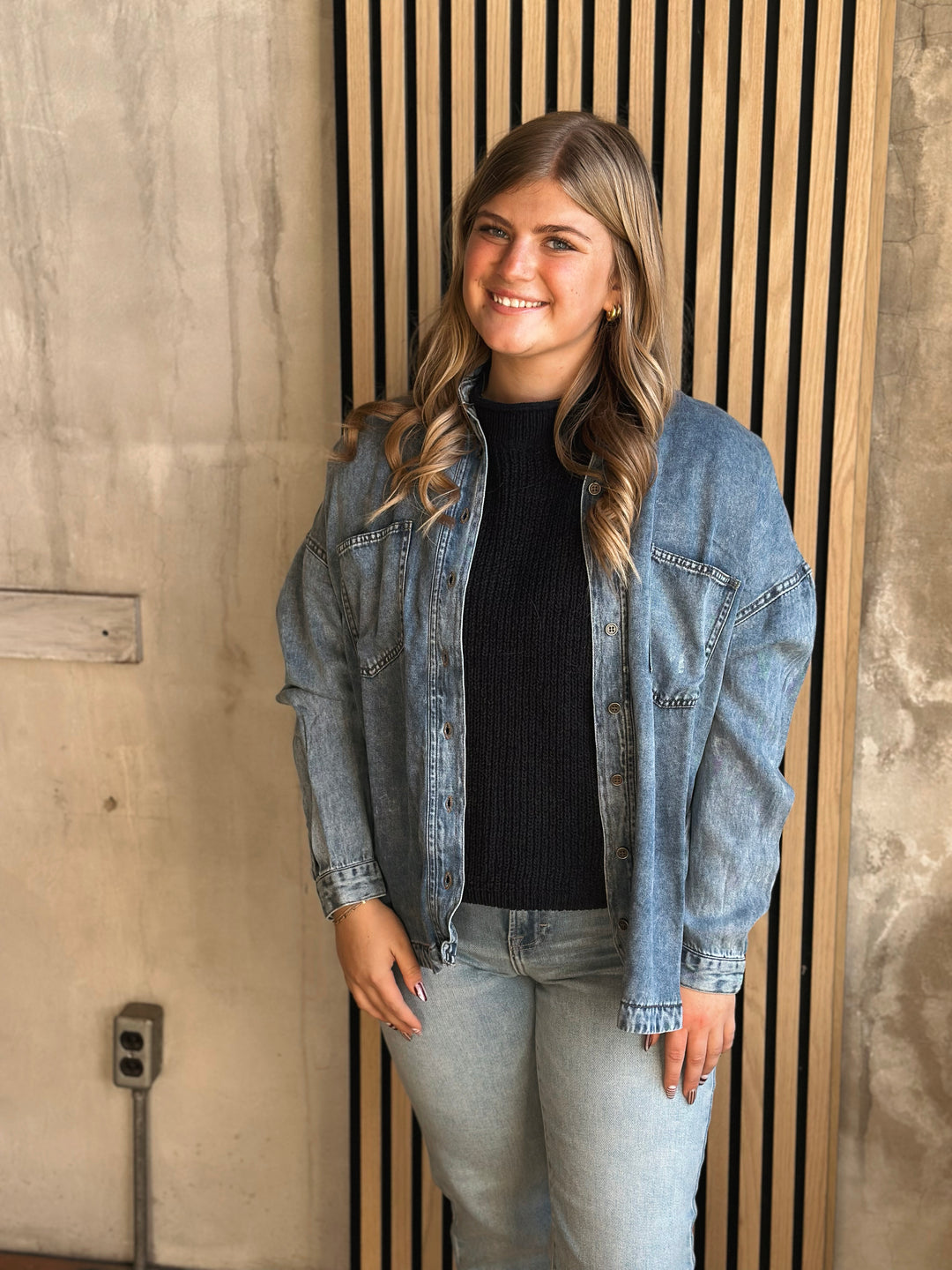 Woman wearing a denim jacket and black shirt standing against a wooden panel wall.