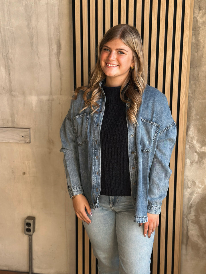 Woman wearing a denim jacket and black shirt standing against a wooden panel wall.