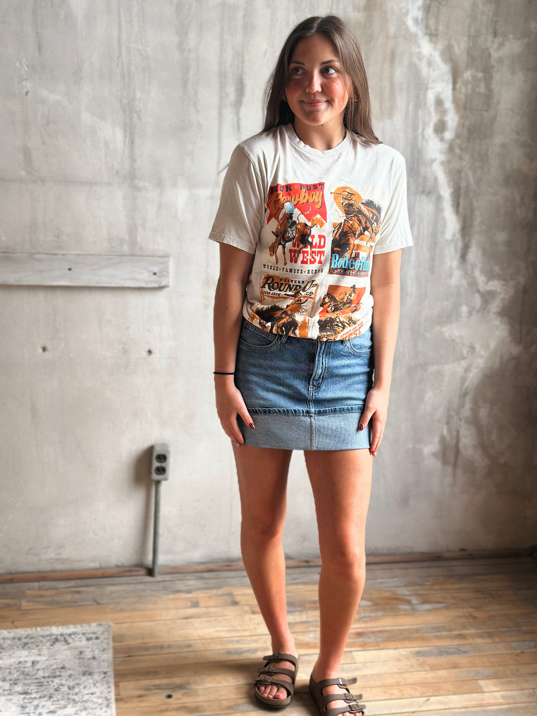 Woman wearing a cowboy themed graphic t-shirt and denim skirt standing against a concrete wall.