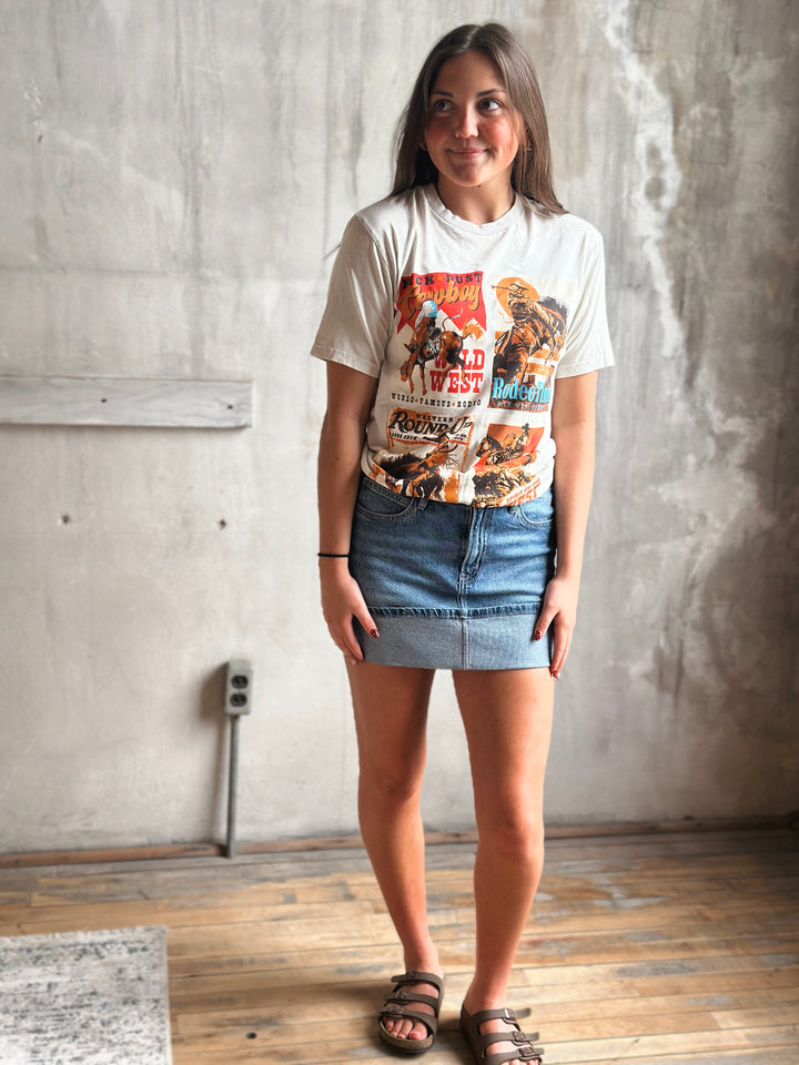 Woman wearing a cowboy themed graphic t-shirt and denim skirt standing against a concrete wall.