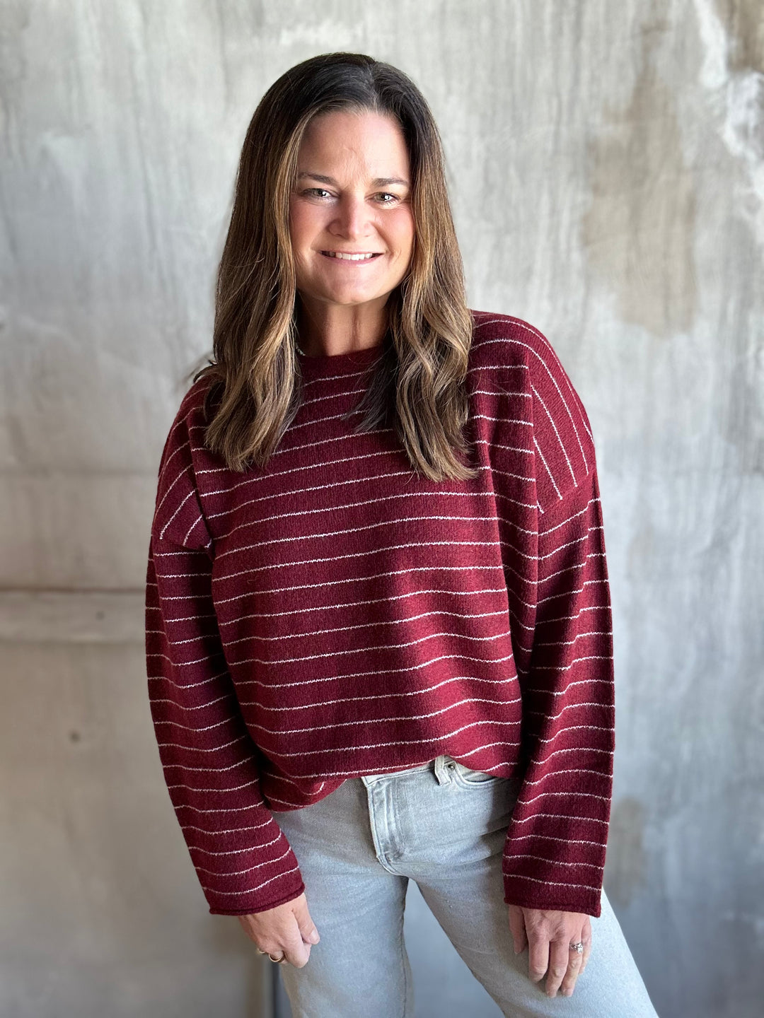 A woman wearing a maroon long-sleeve knit sweater with thin white horizontal stripes and light-wash jeans, standing against a textured gray concrete wall.