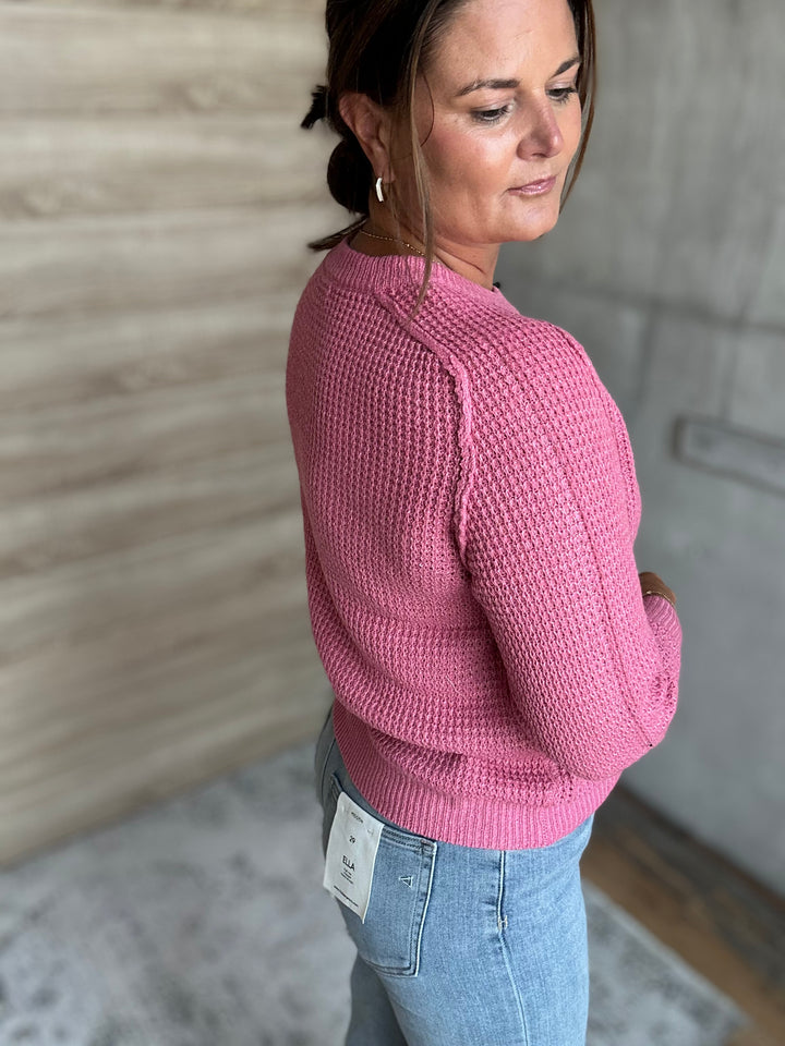 A woman wearing a pink waffle-knit sweater and light blue jeans, shown from a side-back angle in an indoor setting with a wood-paneled wall and concrete floor.