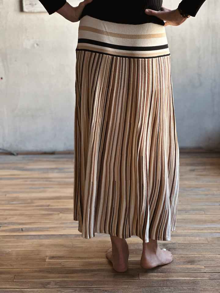 Person wearing a long pleated skirt with a striped pattern on a wooden floor.