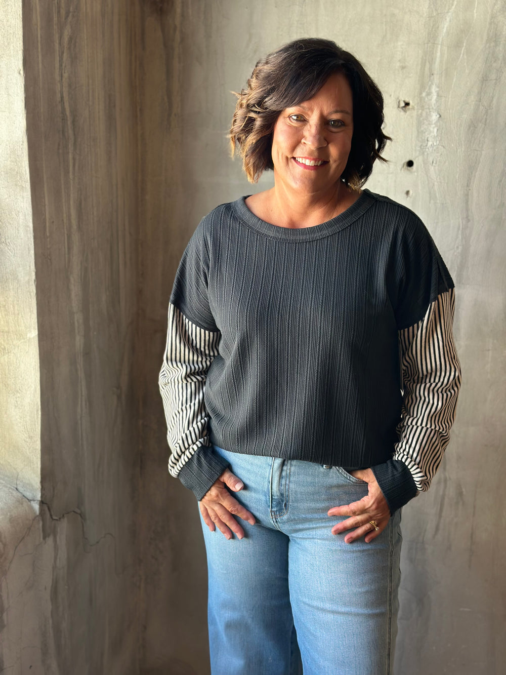 A woman wearing a dark gray ribbed long-sleeve top with striped sleeves and light blue jeans, standing against a textured concrete wall.