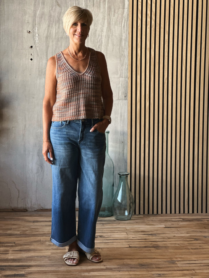 Woman wearing a plaid tank top and blue jeans standing in a room with wooden flooring and a textured wall.