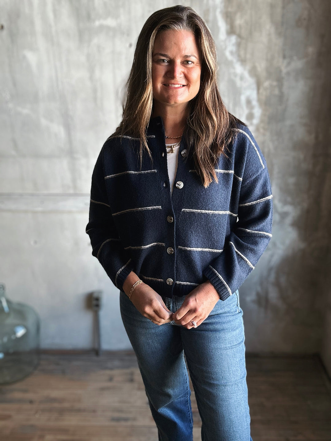 Woman wearing a blue striped cardigan and jeans standing against a textured wall.