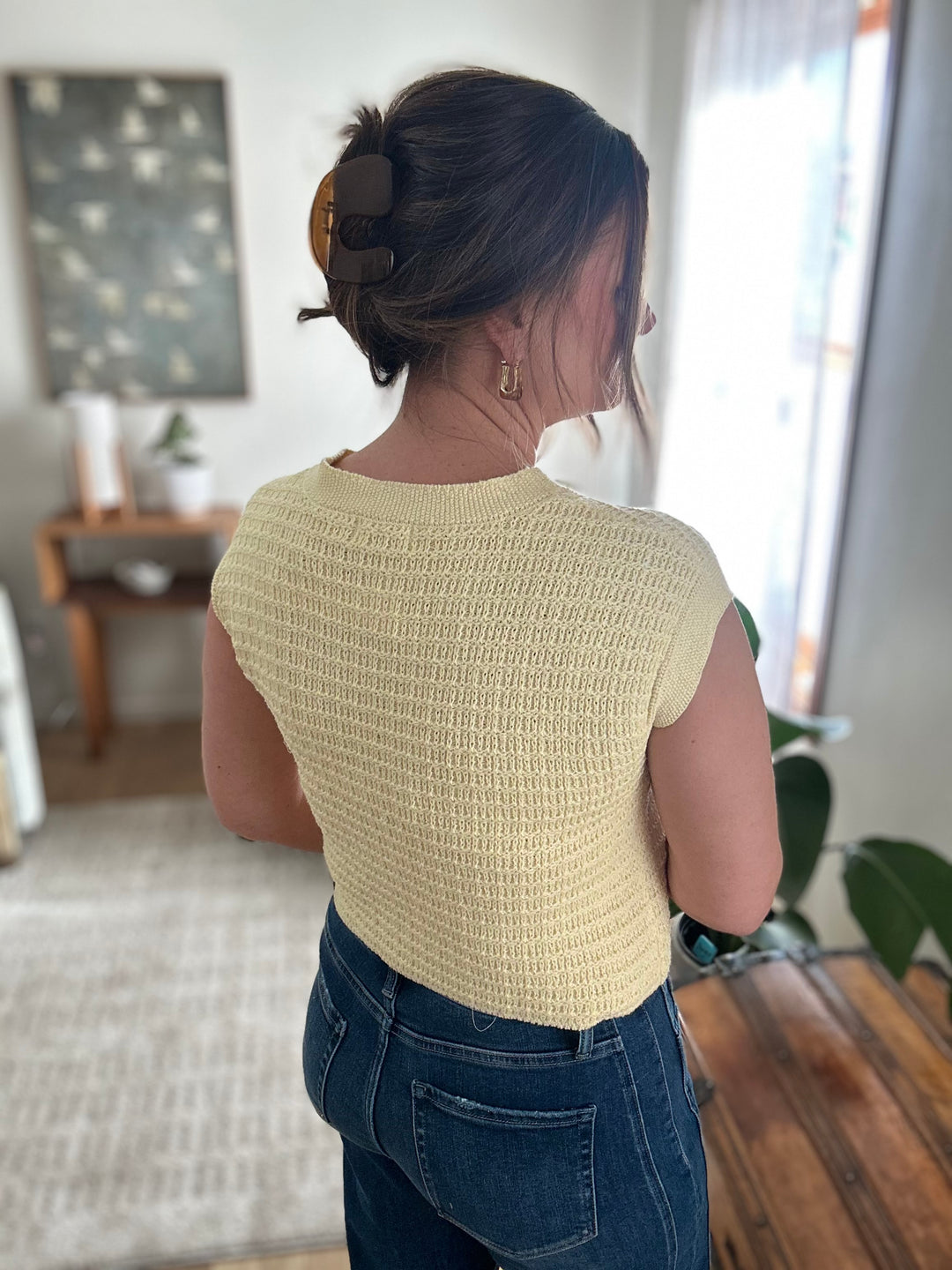 Person wearing a yellow knitted top and blue jeans in a room with a window and furniture.