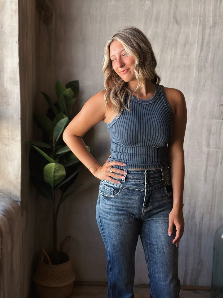 Woman wearing a blue ribbed tank top and jeans standing indoors with a plant in the background.