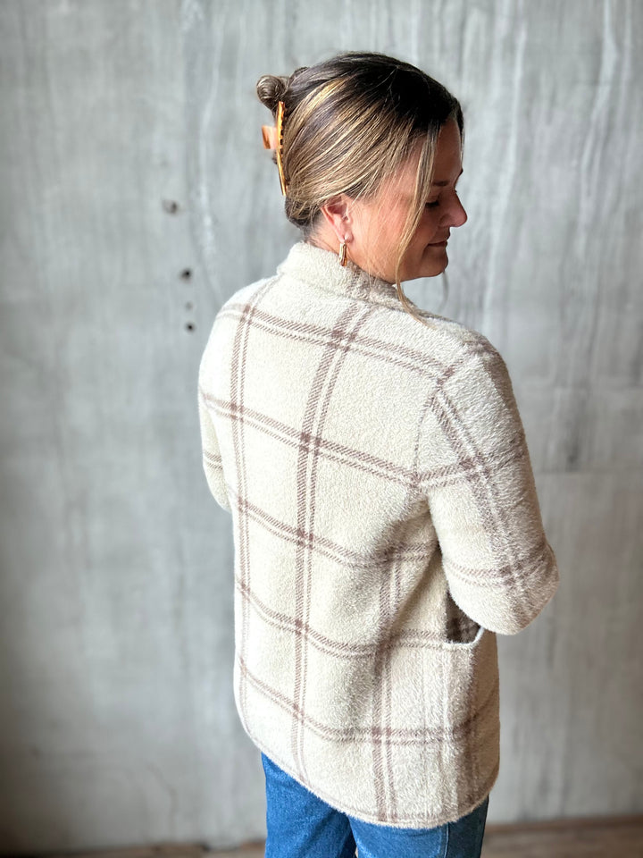 A woman wearing a cream-colored, high-neck, long-sleeve fleece jacket with a brown plaid pattern, paired with blue jeans, standing against a textured gray concrete wall.