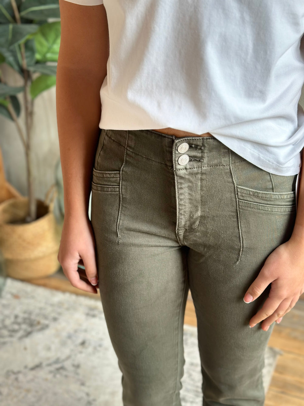 A person wearing olive green button-front jeans with a white t-shirt, shown from the chest down in an indoor setting with a potted plant in the background.