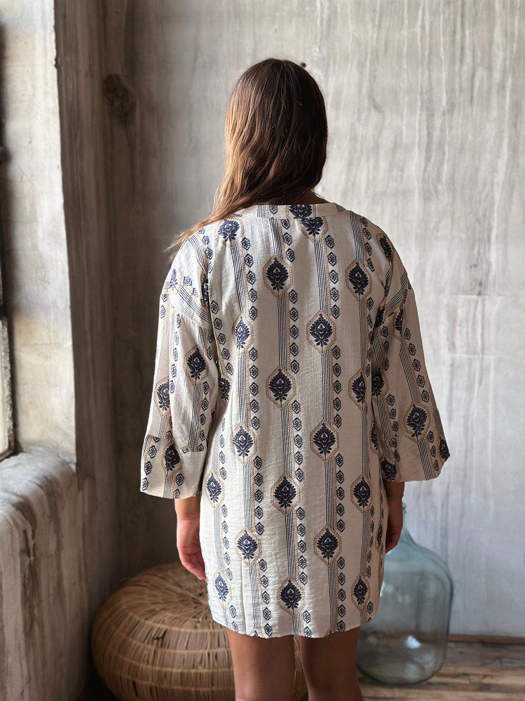 A woman wearing a patterned dress with long sleeves in a room with wooden walls and a large vase.