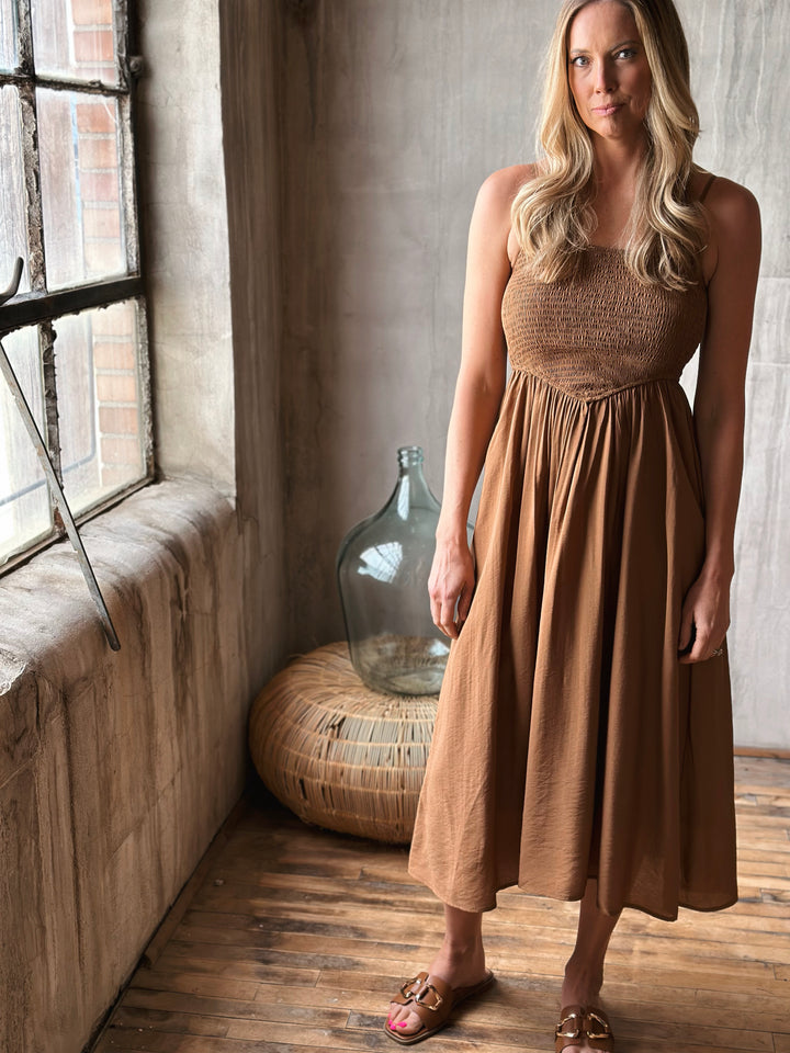 Woman in a brown dress standing in a room with wooden floor and large window.