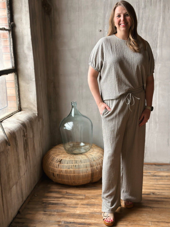 Woman in a textured sage outfit standing in a room with wooden floor and large glass container.
