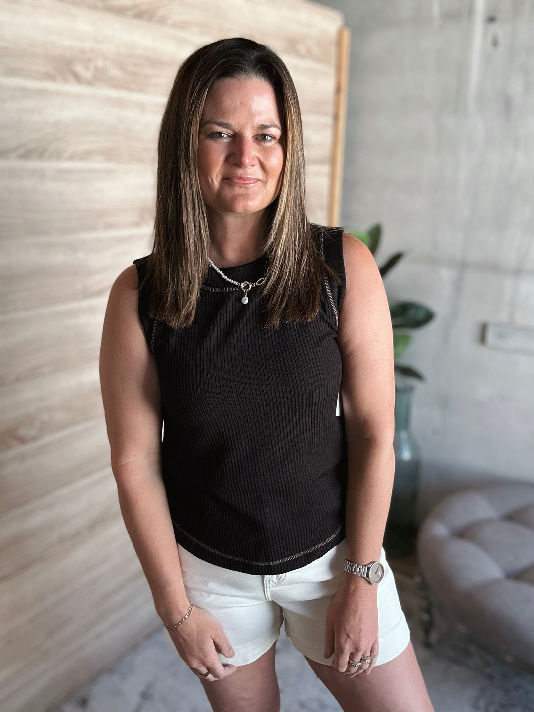 Woman wearing a black sleeveless top with a viable seam around the neck line, shoulders, and the bottom of the shirt. Also wearing white denim shorts. 