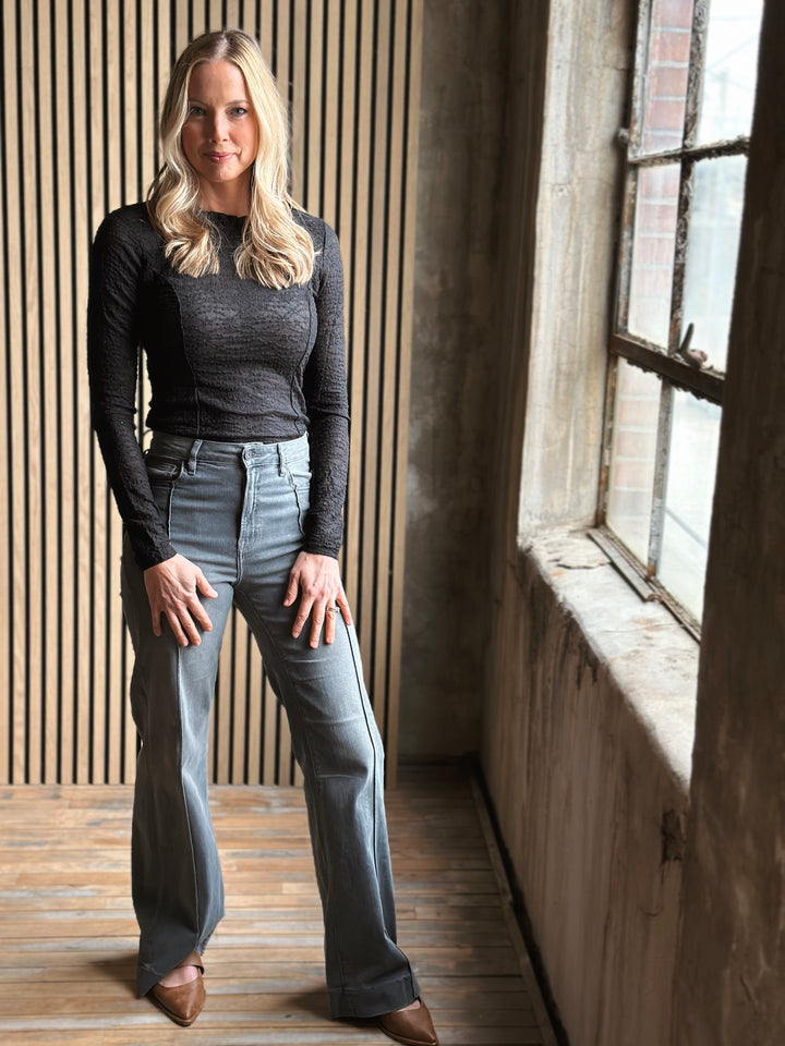 Woman wearing a dark long-sleeve top and light blue jeans standing in a room with wooden floor and large window.