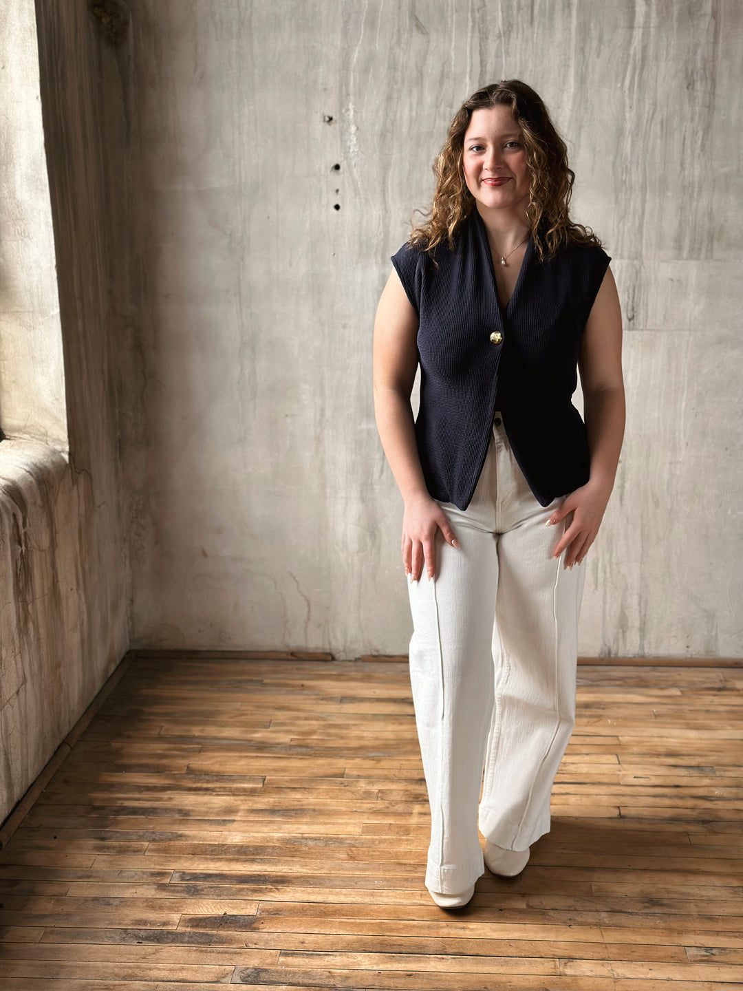 Woman wearing a navy blue sleeveless jacket and white pants standing in a room with wooden floor and concrete wall.