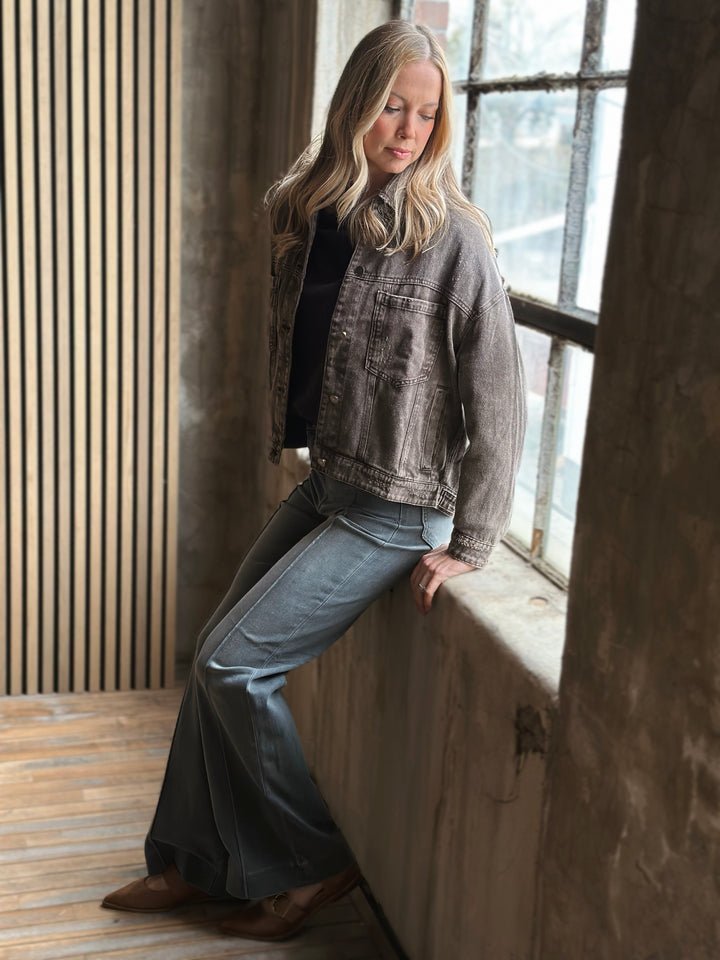 Woman wearing a denim jacket and jeans leaning against a wall indoors.
