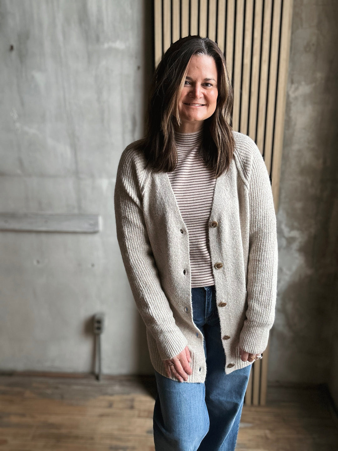 Woman wearing a beige cardigan over a striped shirt with blue jeans, standing against a concrete wall.