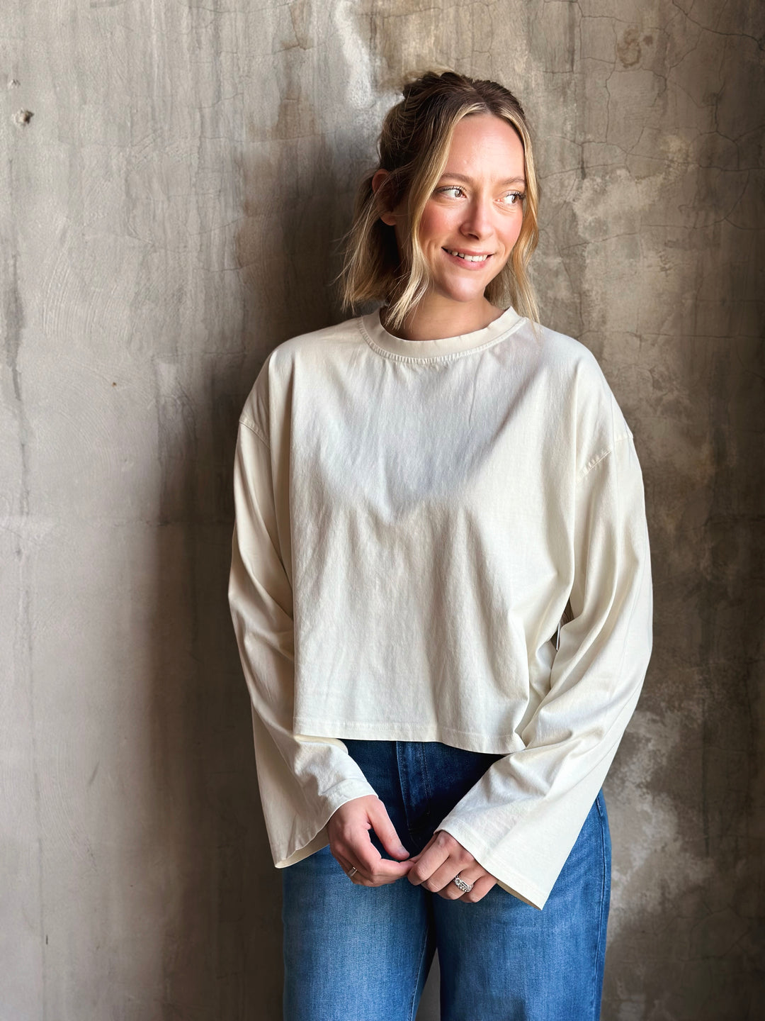 Woman wearing a white long-sleeve top and blue jeans against a textured wall.