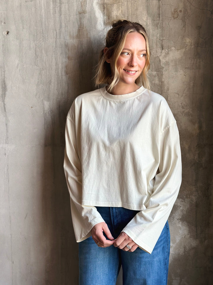 Woman wearing a white long-sleeve top and blue jeans against a textured wall.