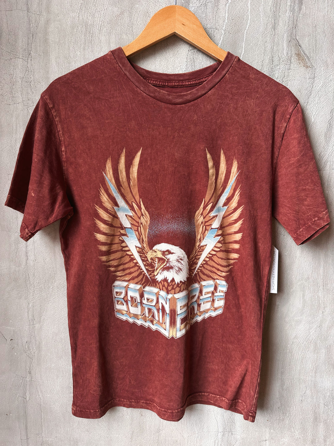 Red t-shirt with eagle graphic and 'Born Free' text on a wooden hanger against a textured wall.