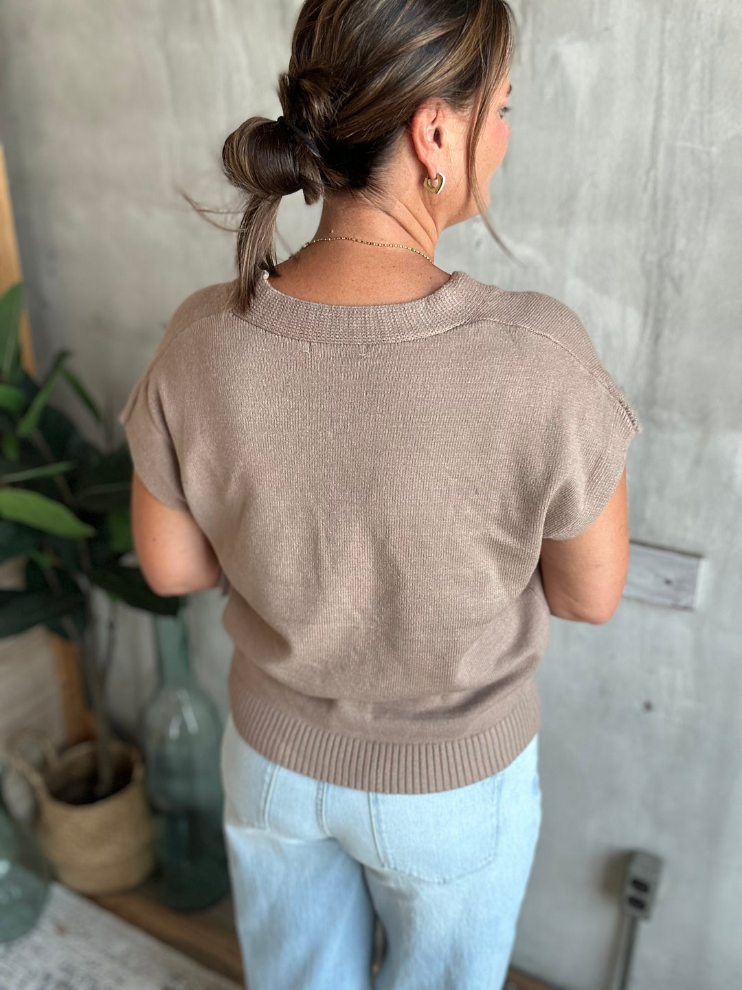 A woman viewed from behind wearing a short-sleeve knit sweater in a warm taupe-brown color, paired with light blue jeans, standing against a textured gray concrete wall.