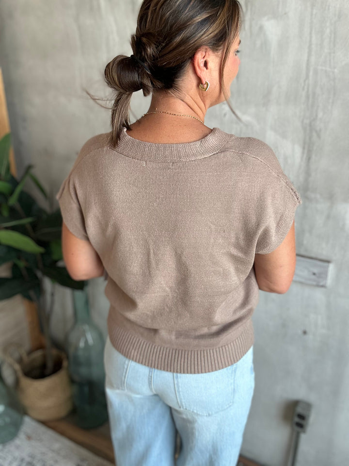 A woman viewed from behind wearing a short-sleeve knit sweater in a warm taupe-brown color, paired with light blue jeans, standing against a textured gray concrete wall.