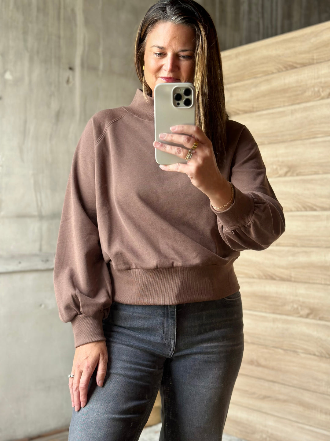 A woman takes a mirror selfie wearing a brown mock neck sweatshirt and dark gray jeans in an indoor setting with a concrete wall and wood paneling.