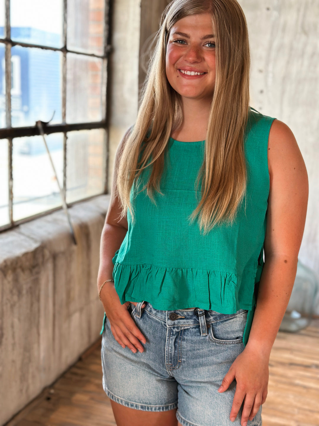 Woman wearing a green sleeveless top and denim shorts indoors.