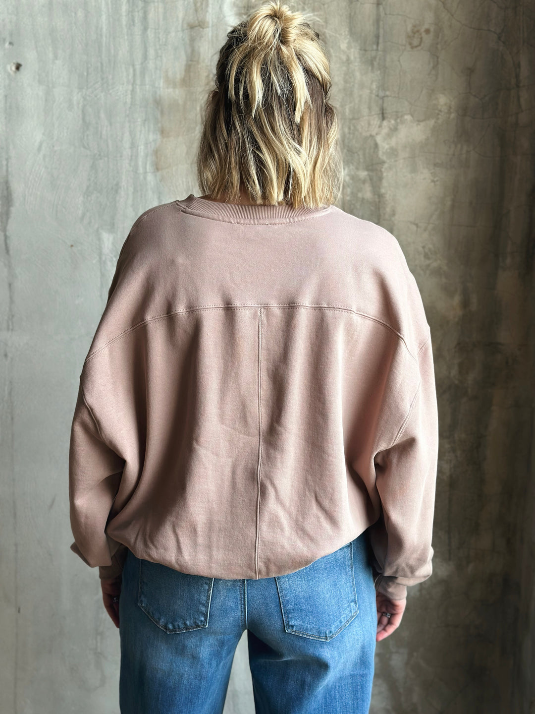 Person wearing a light brown jacket and blue jeans against a textured wall.