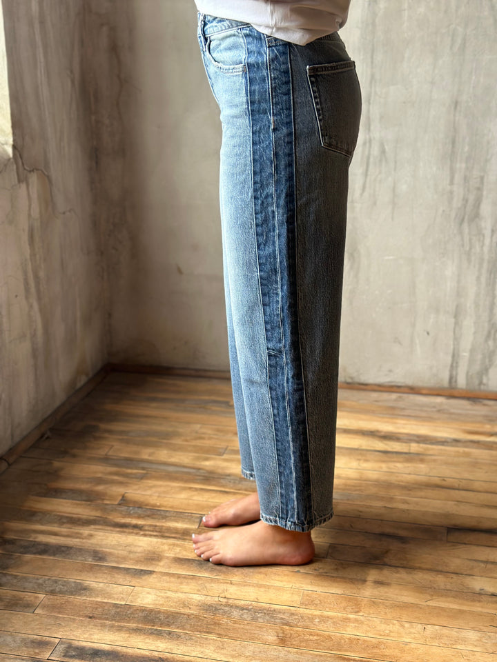 A person wearing light blue denim jeans with a vertical seam detail down the front of the legs, standing barefoot on a wooden floor against a textured concrete wall.