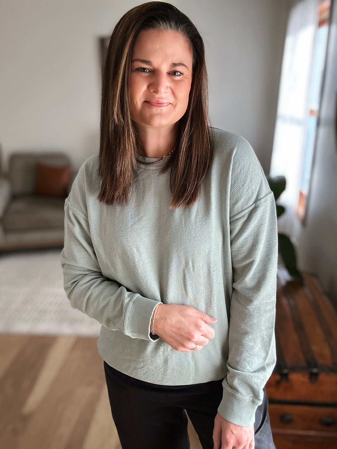 Woman wearing a light gray long-sleeve shirt in a room with a couch and window.