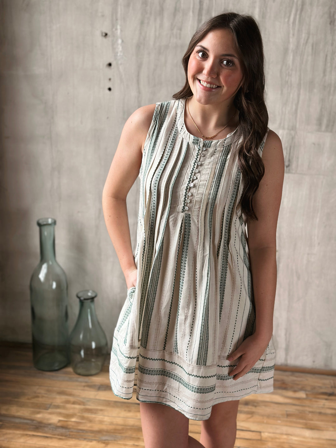 Woman wearing a sleeveless dress with a pattern, standing against a wooden floor and wall.